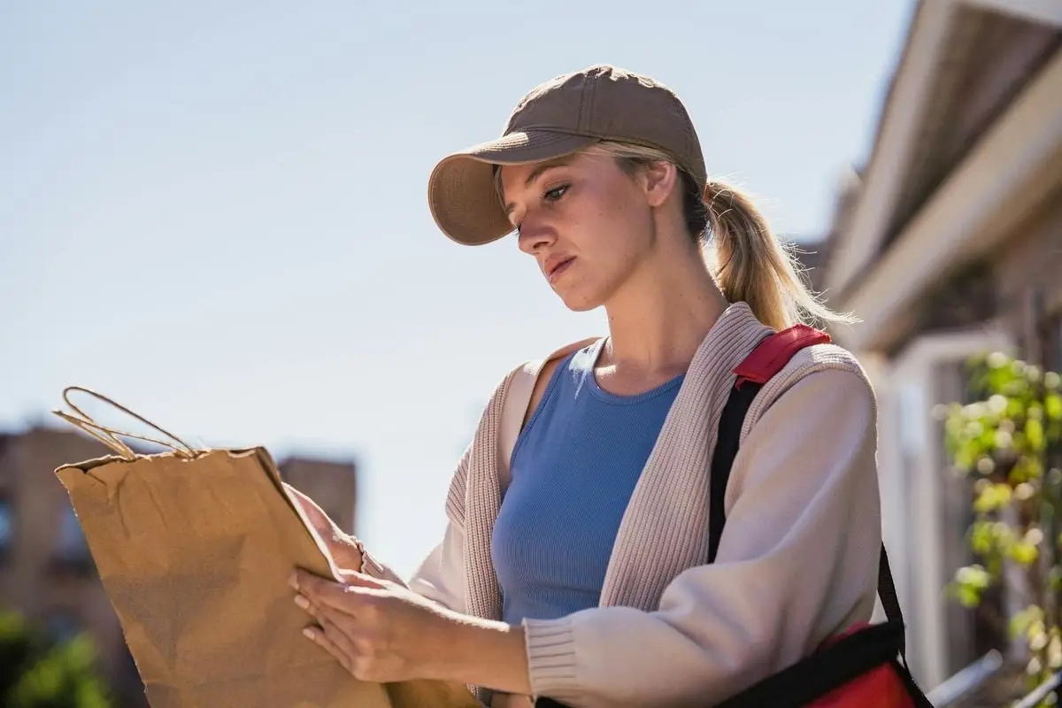 A young woman in casual attire examines a paper bag outdoors on a sunny day.