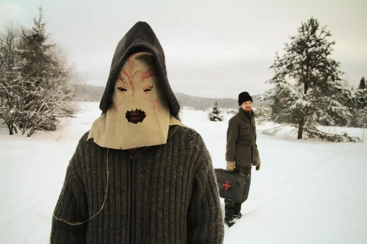 Anonymous man in fabric pagan mask standing against cheerful man in warm clothes in snowy field with trees in countryside in daytime