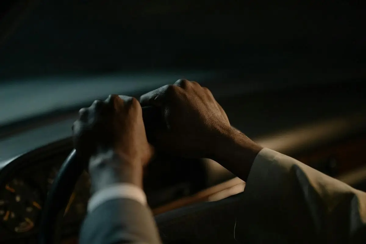 A driver's hands grasp the steering wheel in a dimly-lit vehicle interior, conveying focus and control.