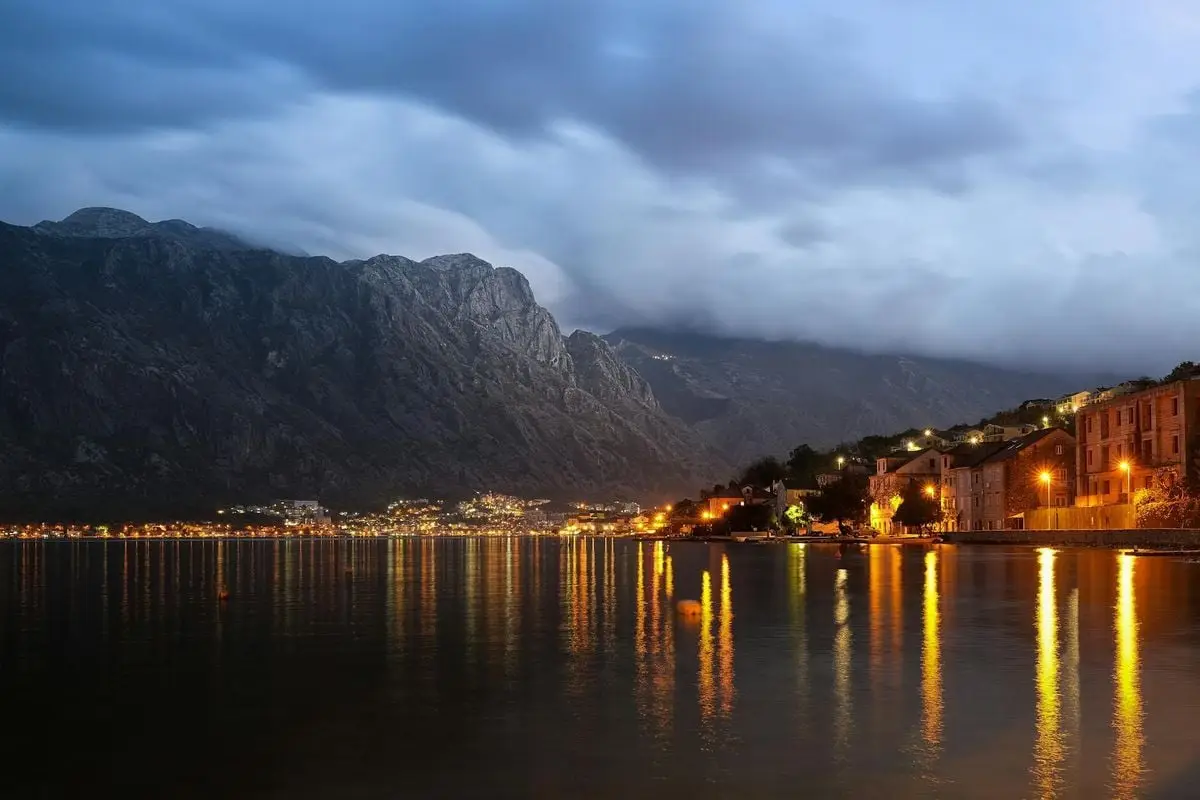 A beautiful night photograph showcasing Kotor's coastline reflecting vibrant lights on the calm sea.