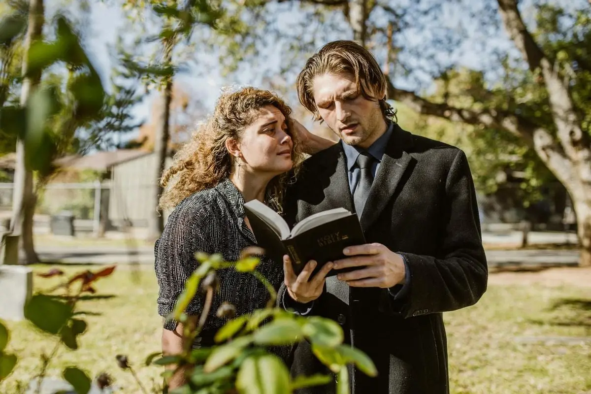 A grieving man and woman read the Bible together in a cemetery setting.