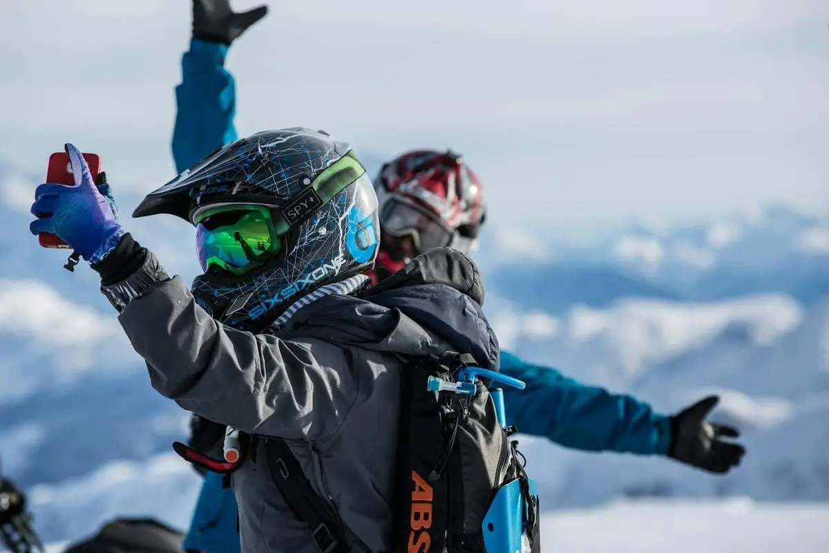 Two snowmobile riders wearing helmets take a selfie amidst snowy mountain peaks.