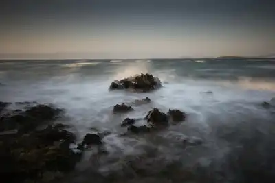 Misty coastal scene at dawn, capturing waves crashing on rocks with a tranquil horizon.