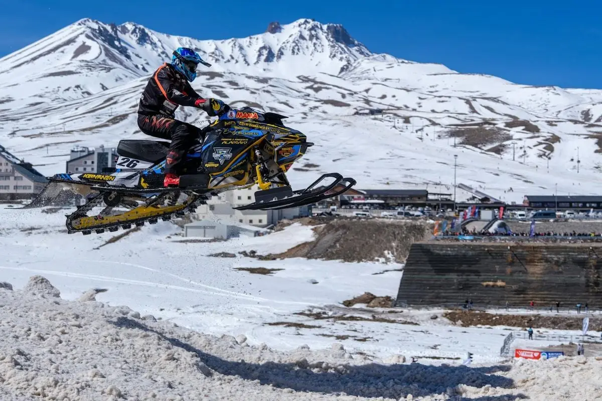 Snowmobiler performing a high jump at a snowy mountain resort with ski slopes and clear blue sky.