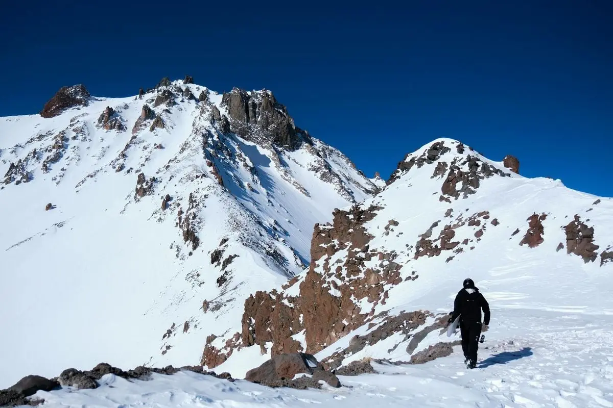 A hiker dressed in black ascends the snow-covered slopes of Mount Erciyes under a clear blue sky.