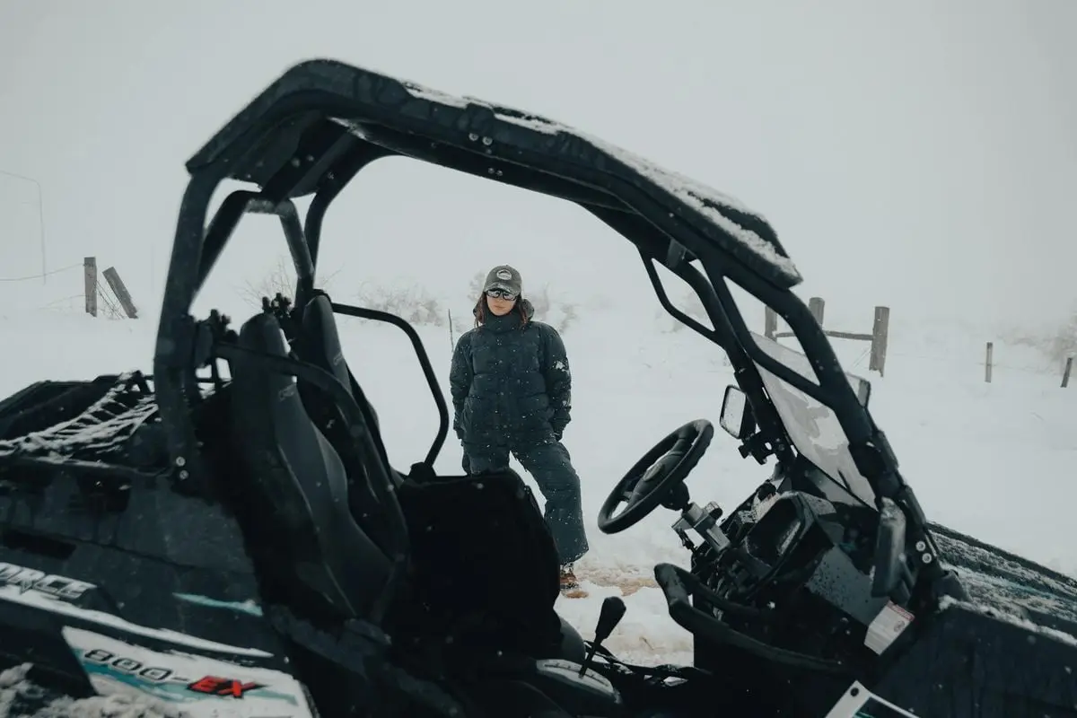 A woman stands in snowy landscape beside an off-road vehicle, wearing winter clothing and sunglasses.