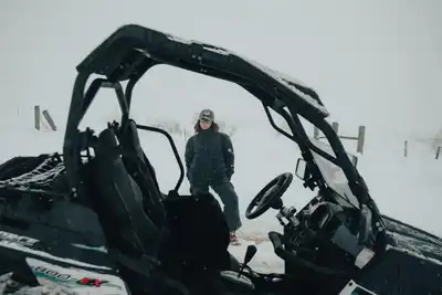 A woman stands in snowy landscape beside an off-road vehicle, wearing winter clothing and sunglasses.