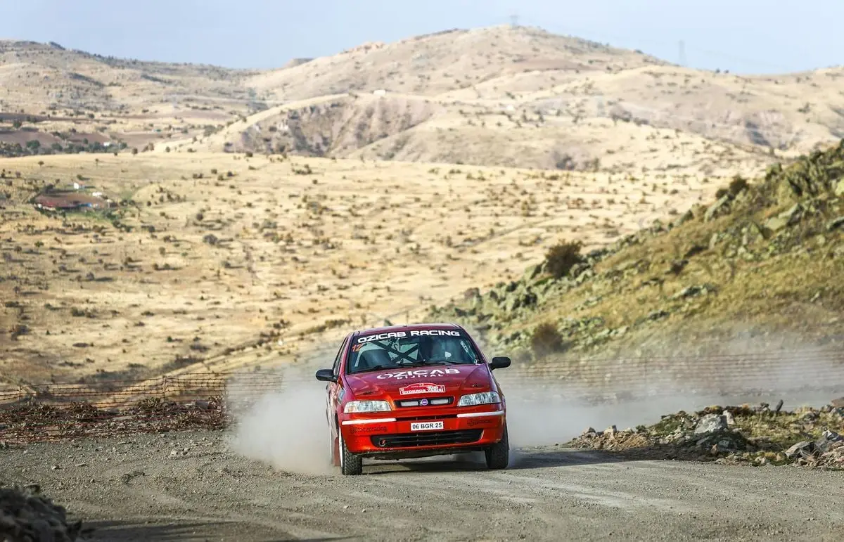 Red rally car speeding through a dusty desert landscape, leaving a trail of dust behind.