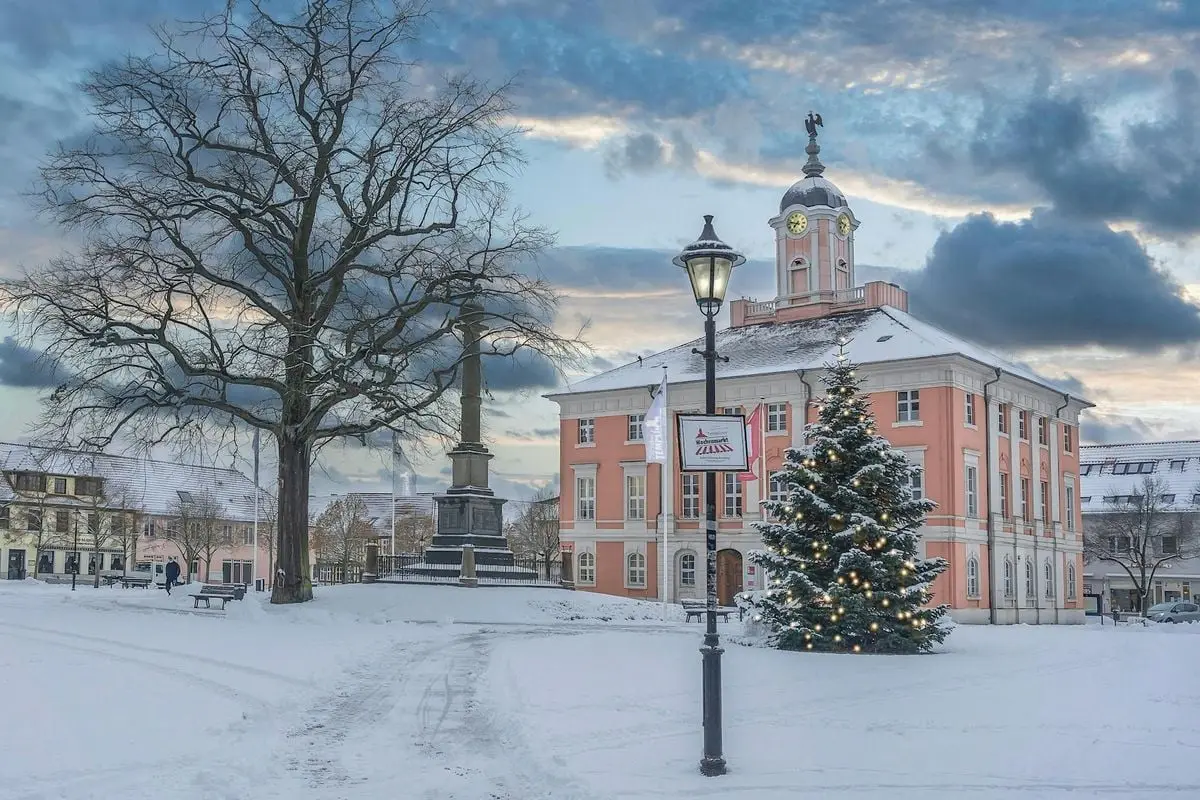 Snowy town square with Christmas tree and historic architecture under evening sky.