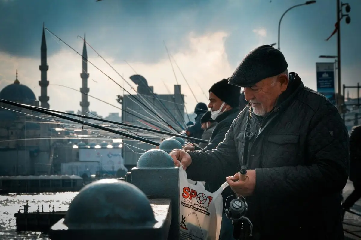 An elderly man enjoys fishing on a lively bridge in Istanbul with mosques in the background.