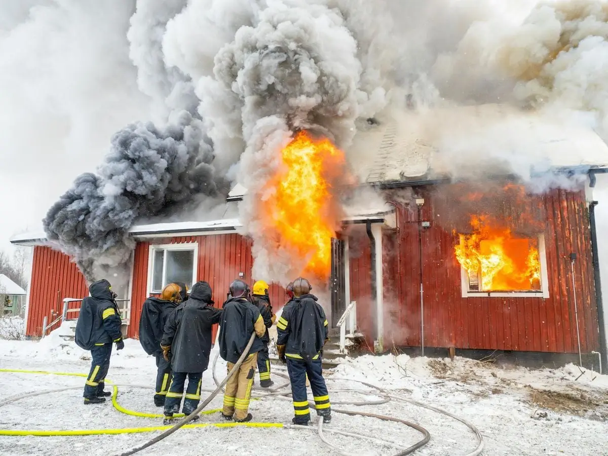 Firefighters combat a house fire amidst heavy smoke and flames in a snowy setting.