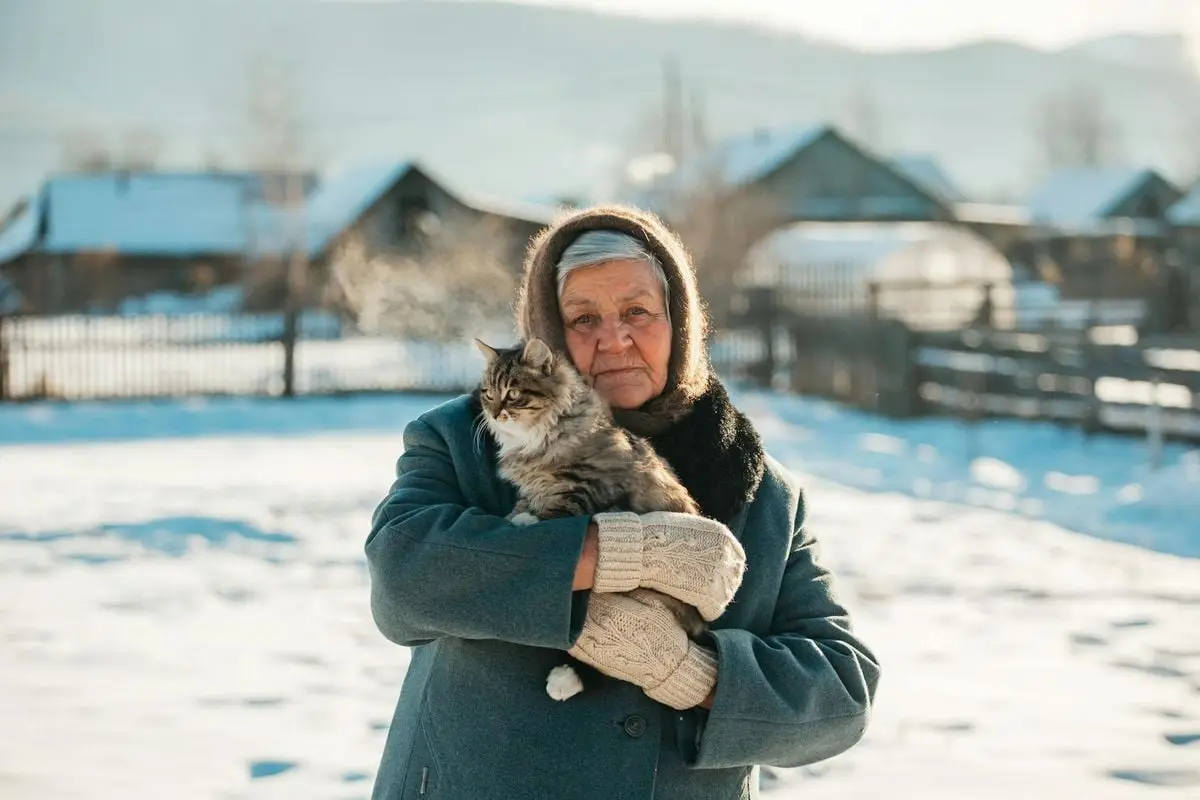 Senior woman holding a fluffy cat in a snowy village setting during winter.