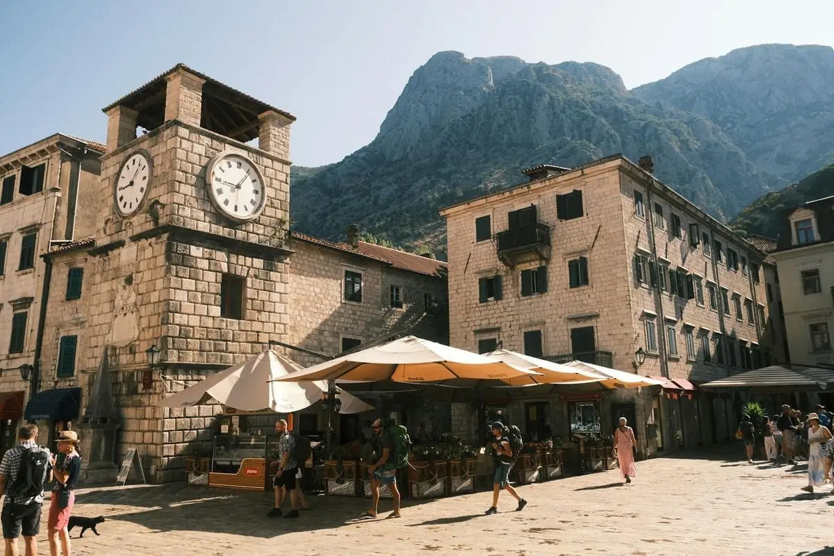 Bustling Kotor old town square with historic clock tower and stone buildings, framed by mountains.