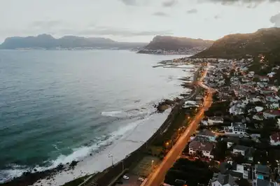 Drone view of a picturesque coastal city at twilight with mountains and illuminated streets.