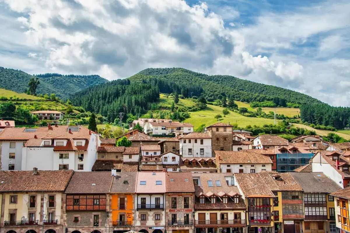Charming town with terracotta roofs against a lush green mountainous backdrop under a vibrant sky.