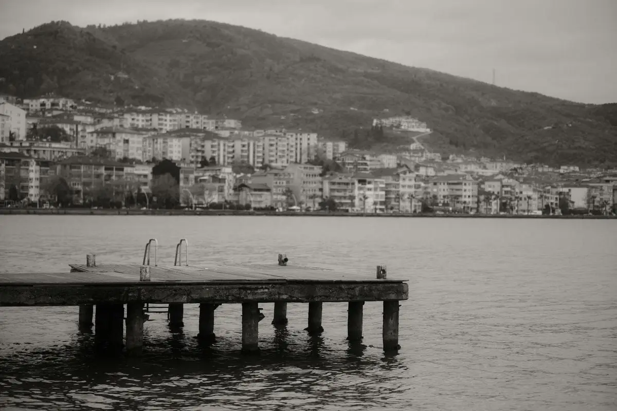 Black and white photo of Degirmendere dock, tranquil waters, cityscape, and hills.
