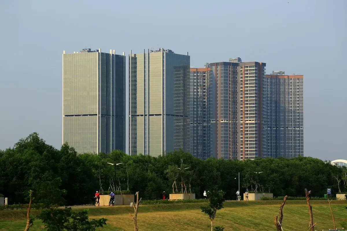 Tall modern buildings towering over a lush green park with walking paths and people.