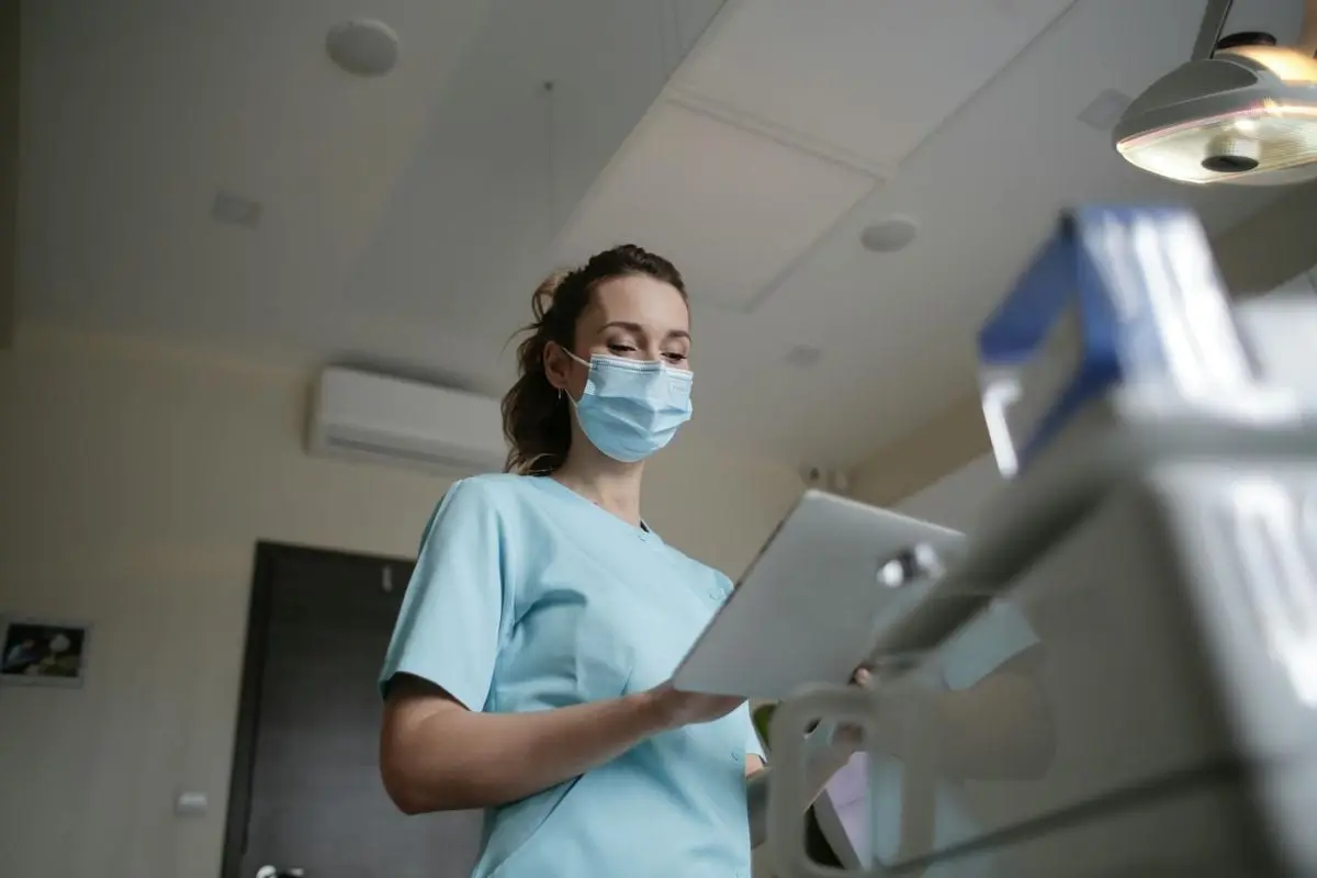Female healthcare worker wearing a mask and holding a clipboard in a clinical environment.