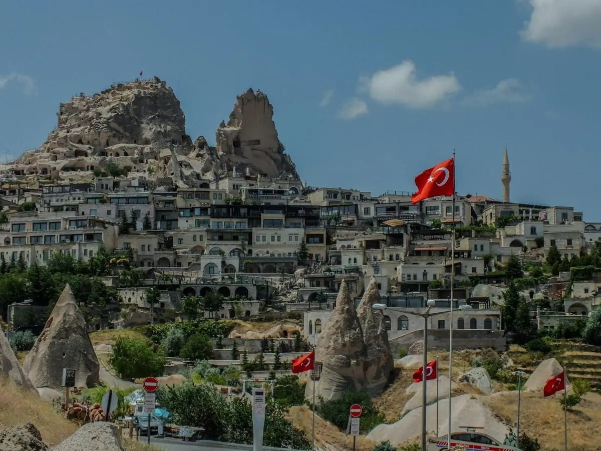 Scenic view of Uchisar Castle in Turkey with Turkish flags in foreground, under a clear blue sky.