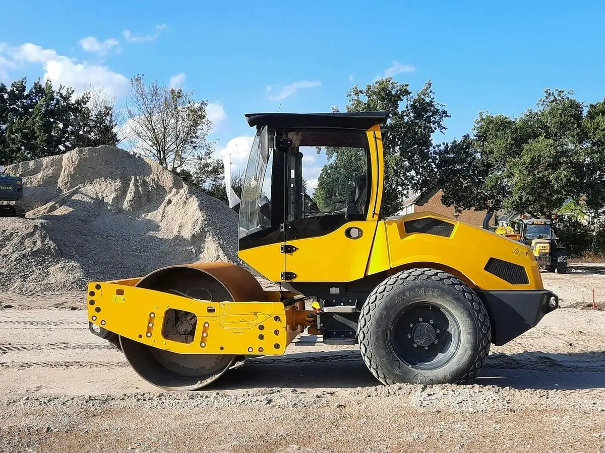 Close-up of a yellow road roller on a construction site with gravel and trees in the background.