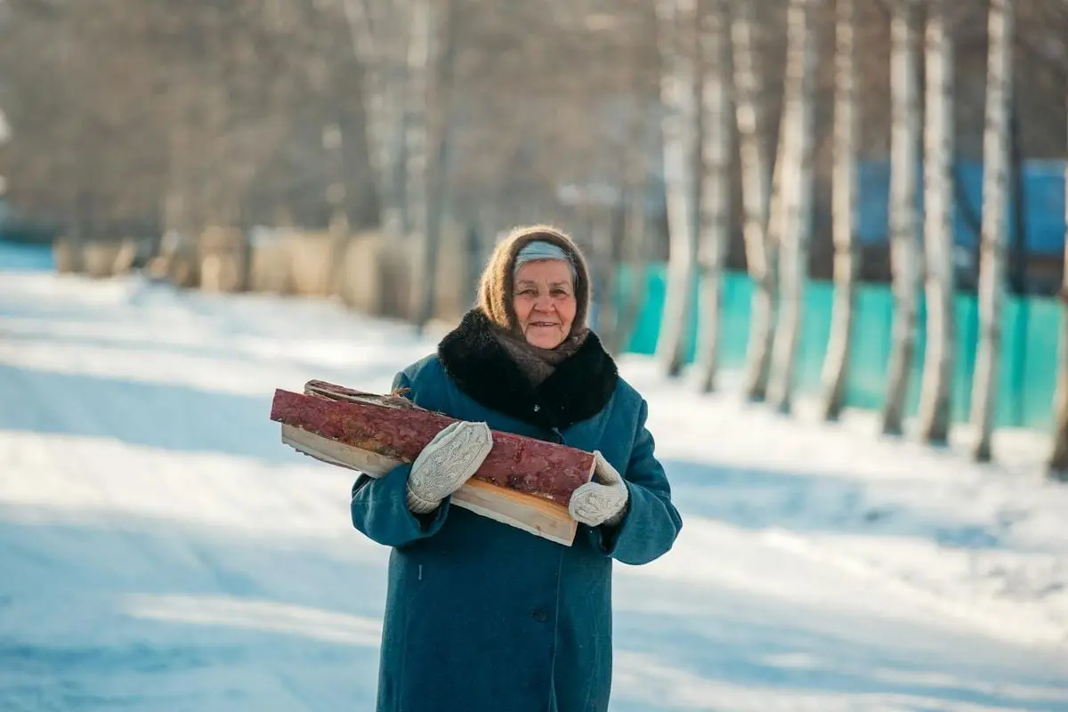 Senior woman in winter clothes carrying logs, outdoors in a snowy landscape.