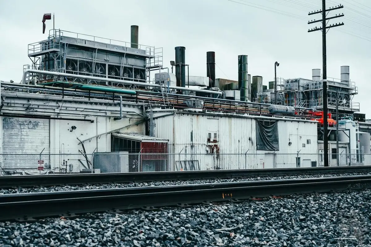 A weathered factory building with visible pipelines and railway tracks under an overcast sky.
