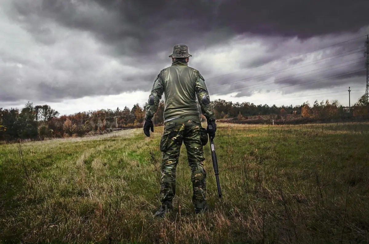 A soldier in camouflage uniform with a rifle stands in a grassy field under a cloudy sky.