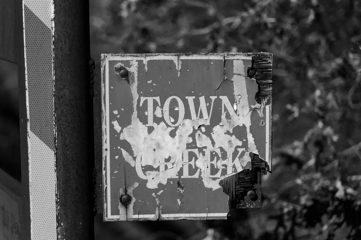 Black and white image of a weathered Town Creek sign outdoors, featuring peeling paint and texture.