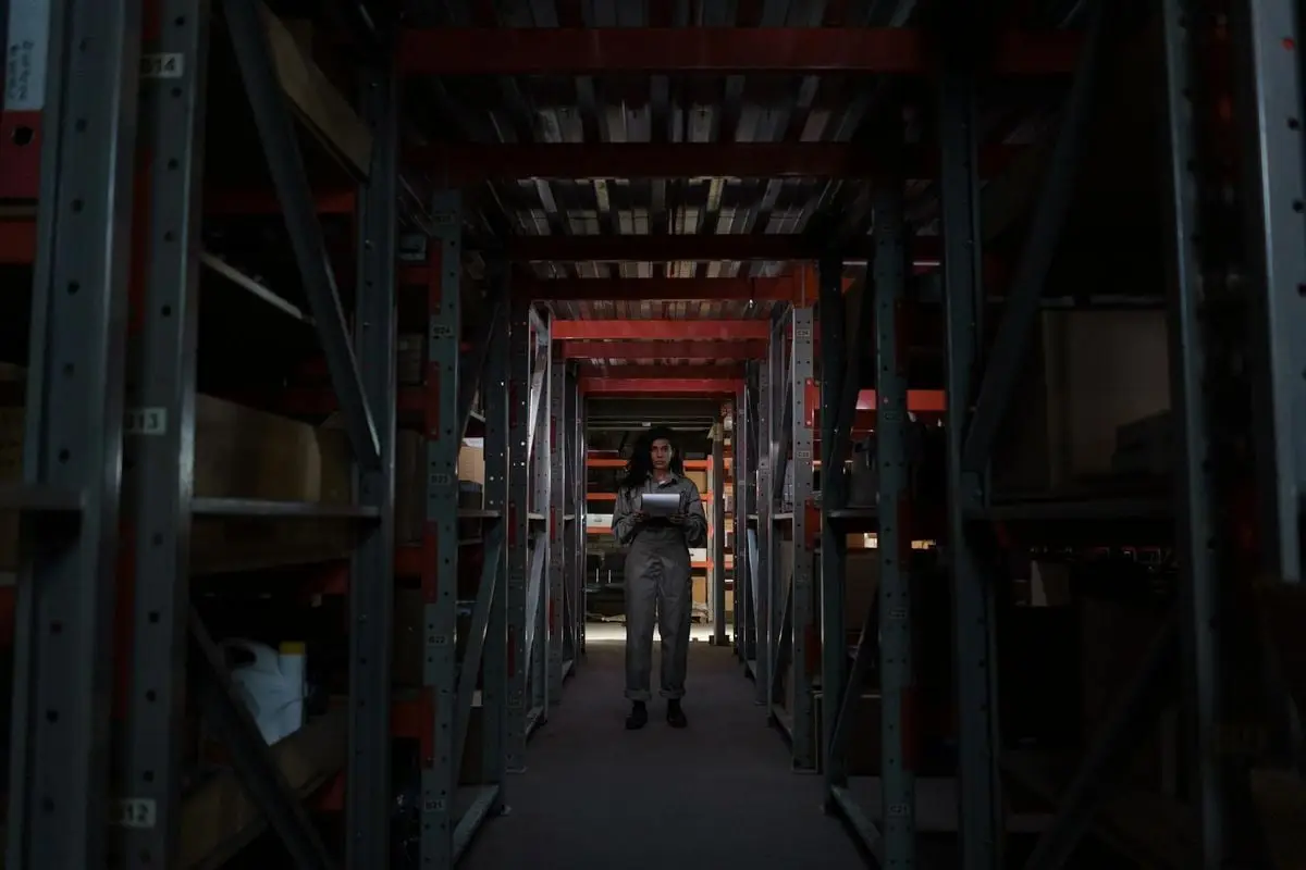 A woman stands in a narrow warehouse corridor, inspecting inventory under dim lighting.