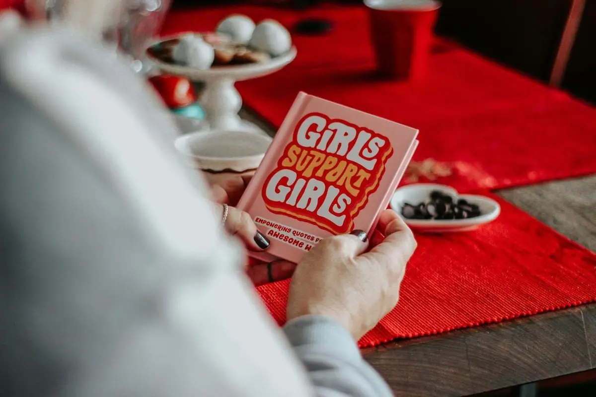 Close-up of a 'Girls Support Girls' book held by hands over a red table setup.
