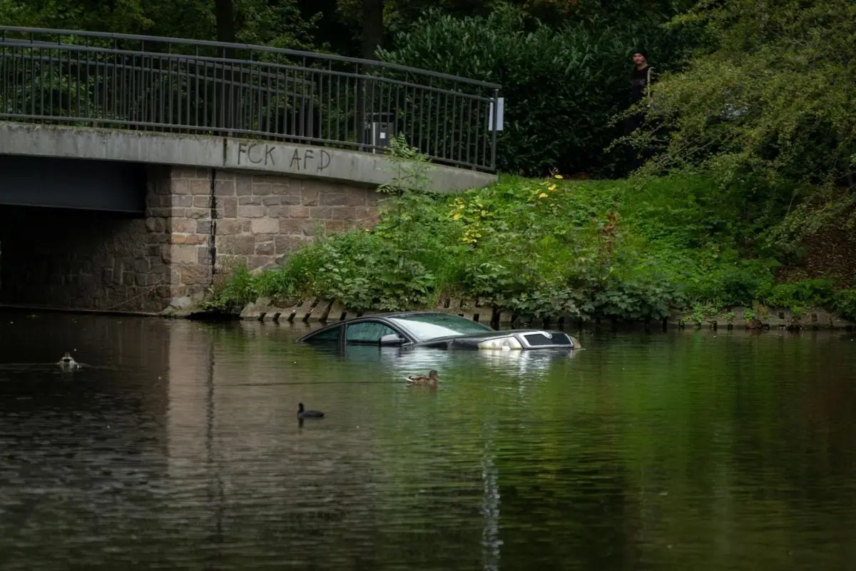 A car submerged in a river near a stone bridge in Chemnitz, Germany, with graffiti and greenery.
