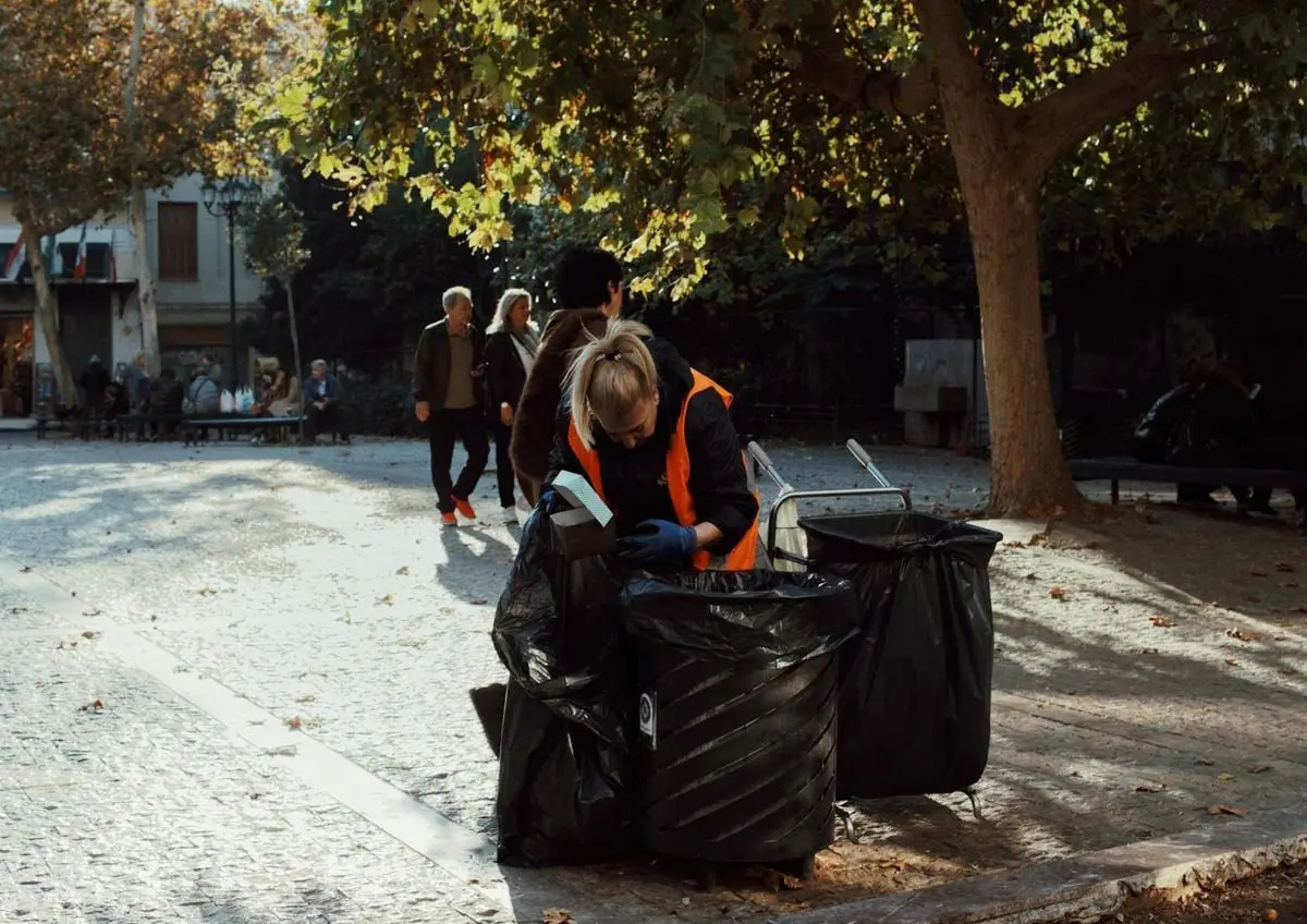 Sanitation worker in orange vest collecting trash in urban park during fall.