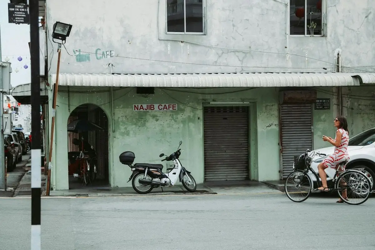 A woman rides past Najib Cafe on a vintage bicycle in an urban setting.