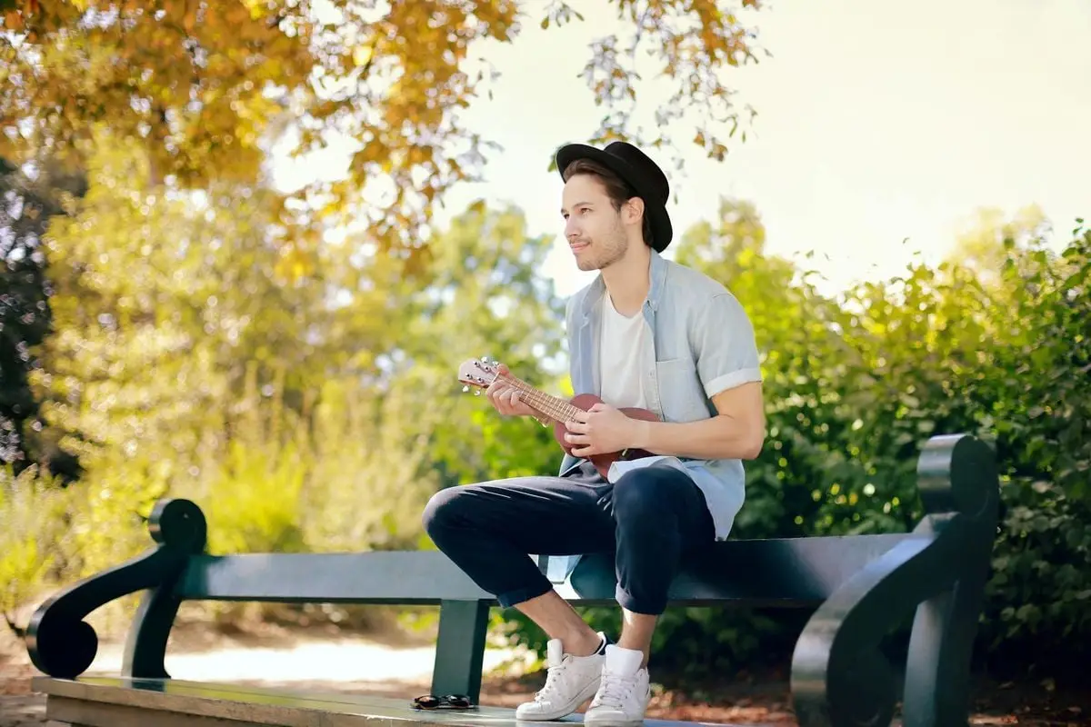 Man enjoying a sunny day playing ukulele on a park bench surrounded by greenery.