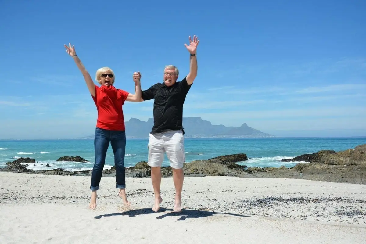 Elderly couple jumping with joy on a beach in Cape Town with Table Mountain in the background.