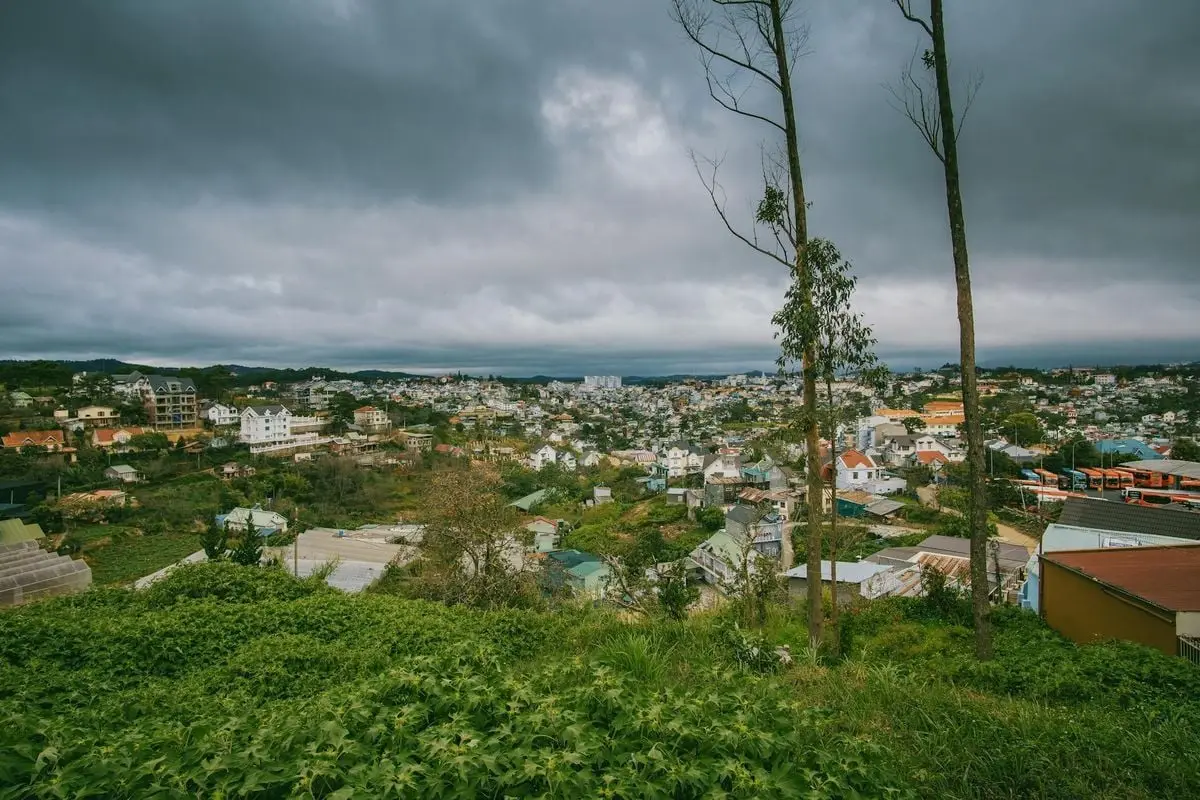 A panoramic view of a city with lush greenery under a dramatic cloudy sky, showcasing architecture and nature.