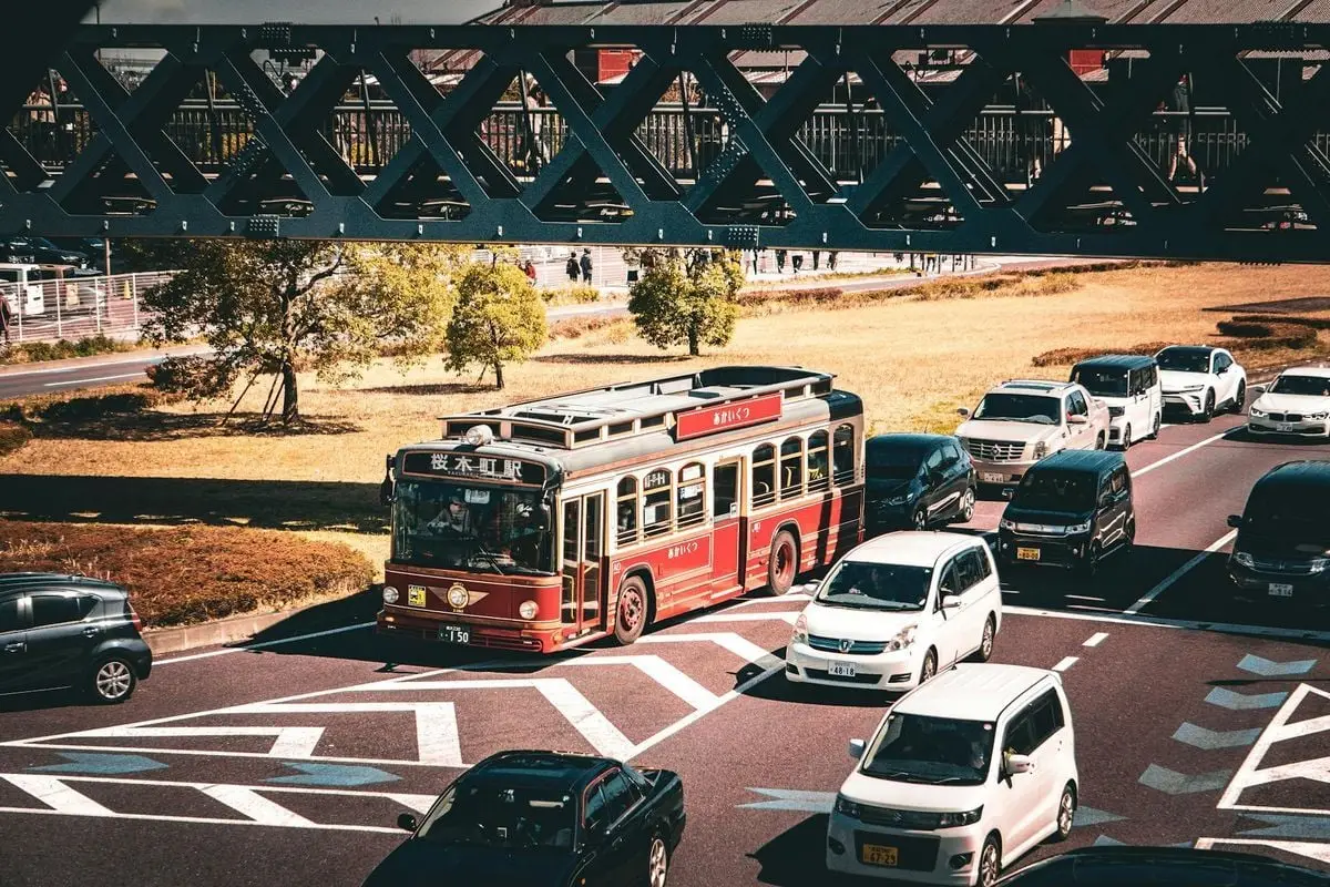 A vintage red bus travels on a busy street under a bridge, surrounded by cars.