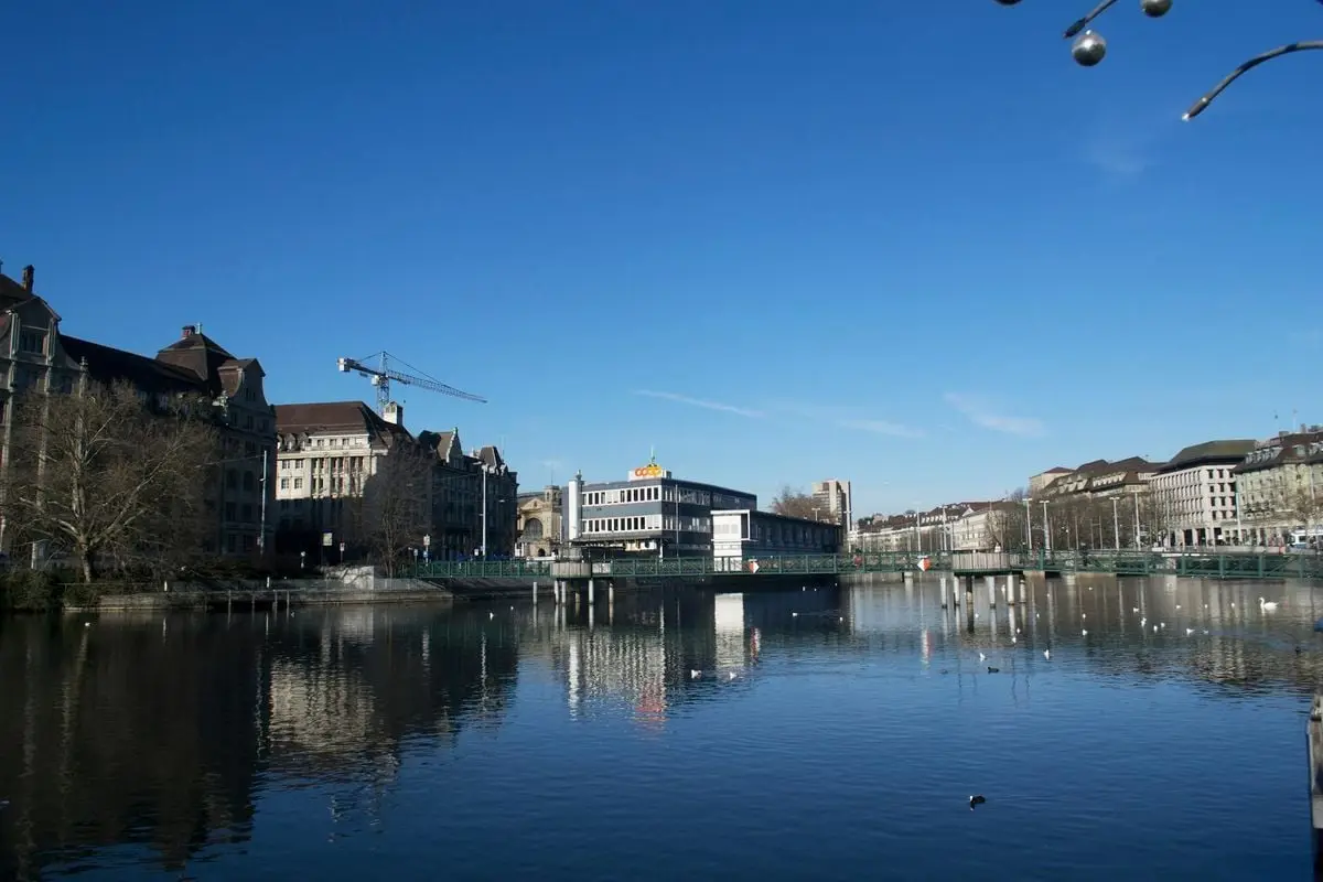 Picturesque scene of Zürich's architecture and waterfront reflecting in the calm river under a clear blue sky.