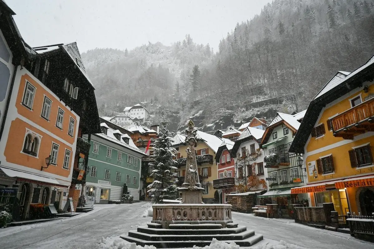 Charming winter view of Hallstatt's colorful town square with snow-covered buildings and a central fountain.