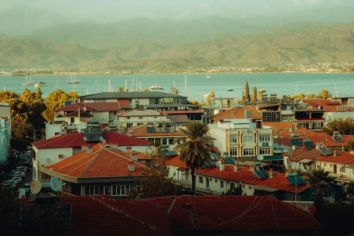 Charming coastal town with red roofs, mountains, and sailboats in the bay under a tranquil sky.