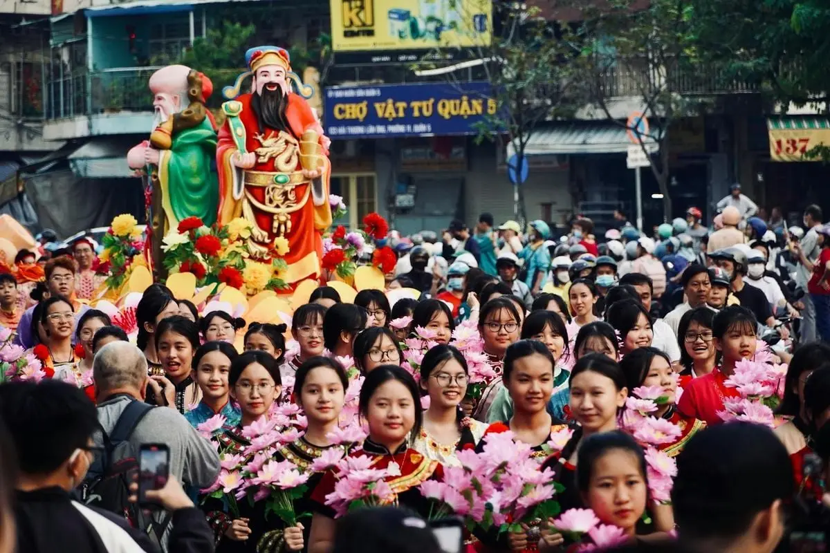 A vibrant street festival parade featuring Asian participants with floral decorations and large statues.