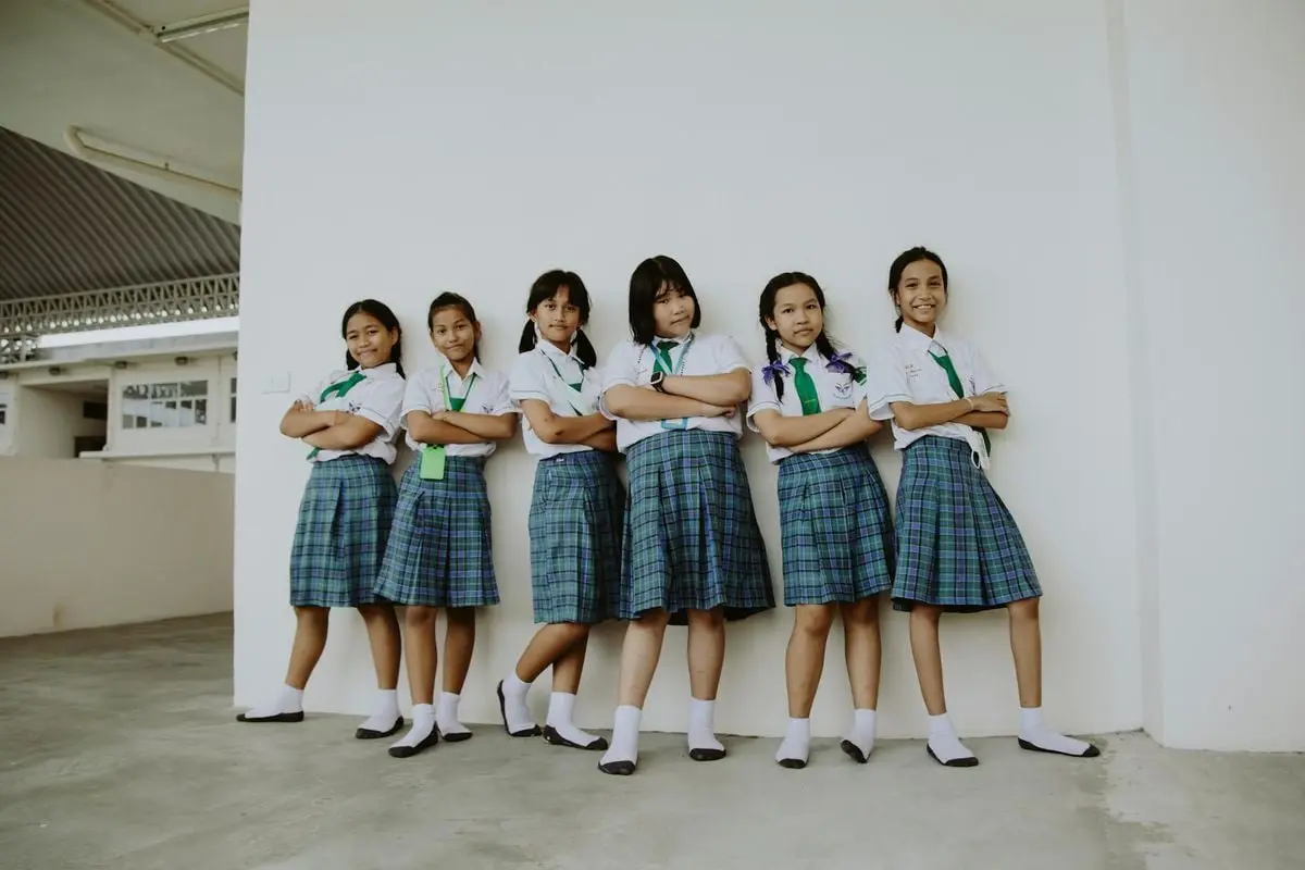 A group of six smiling schoolgirls in uniform standing indoors with arms crossed, displaying friendship.