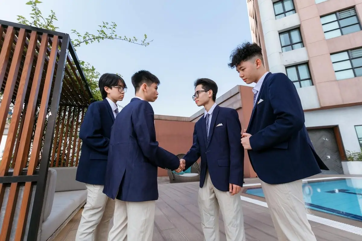 Four teenage boys in school uniforms shaking hands on a rooftop terrace.