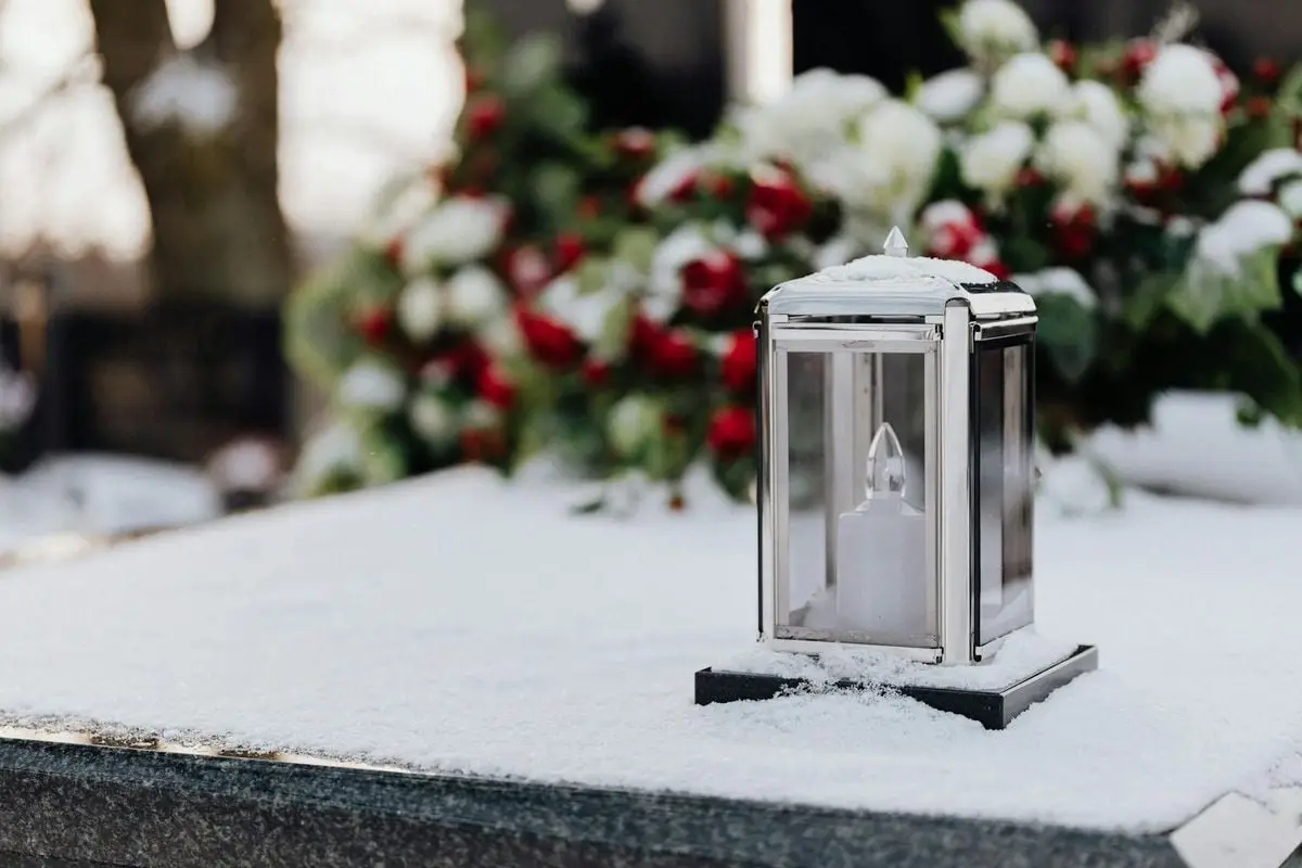 A lantern in snow on a tomb, with blurred colorful flowers in the background.