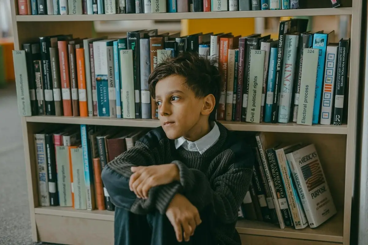 A thoughtful young boy sits alone in a library, surrounded by books.