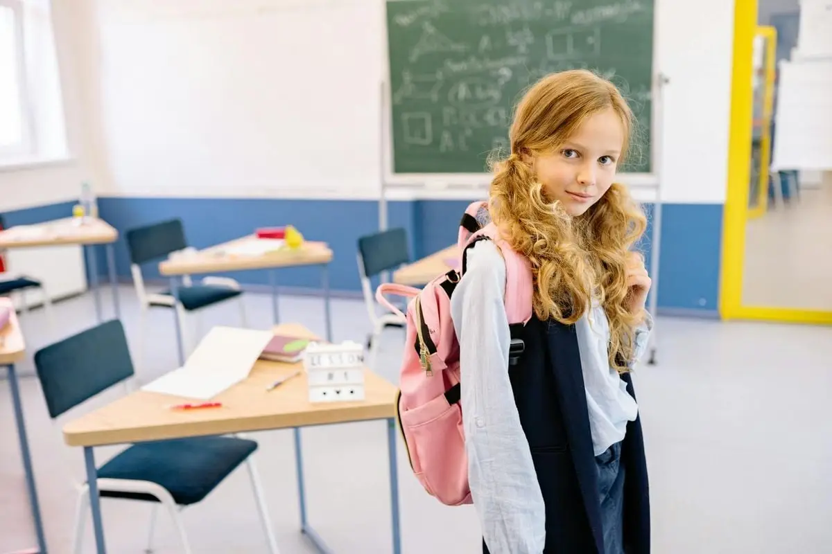 A young girl with a backpack stands confidently in a bright classroom setting.