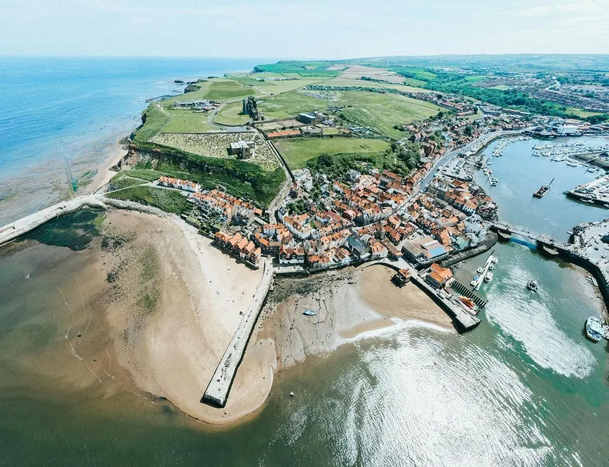 Stunning aerial shot showcasing a coastal town with a pier, beach, and lush green fields under a bright sky.