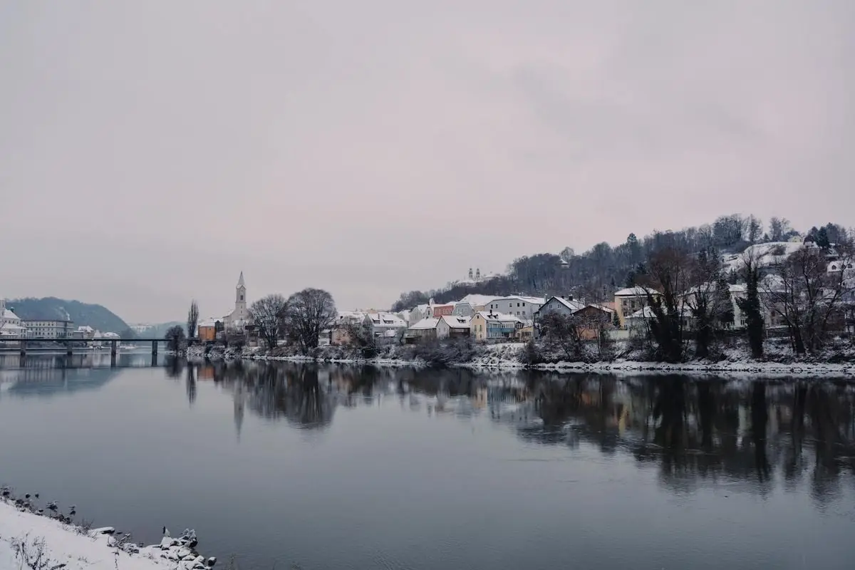 A scenic view of Passavia during winter with snow-covered houses reflecting on the river.