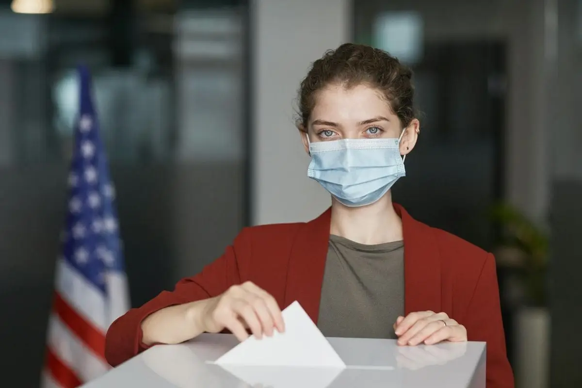 A woman wearing a face mask casts her vote in a ballot box on Election Day with an American flag in the background.