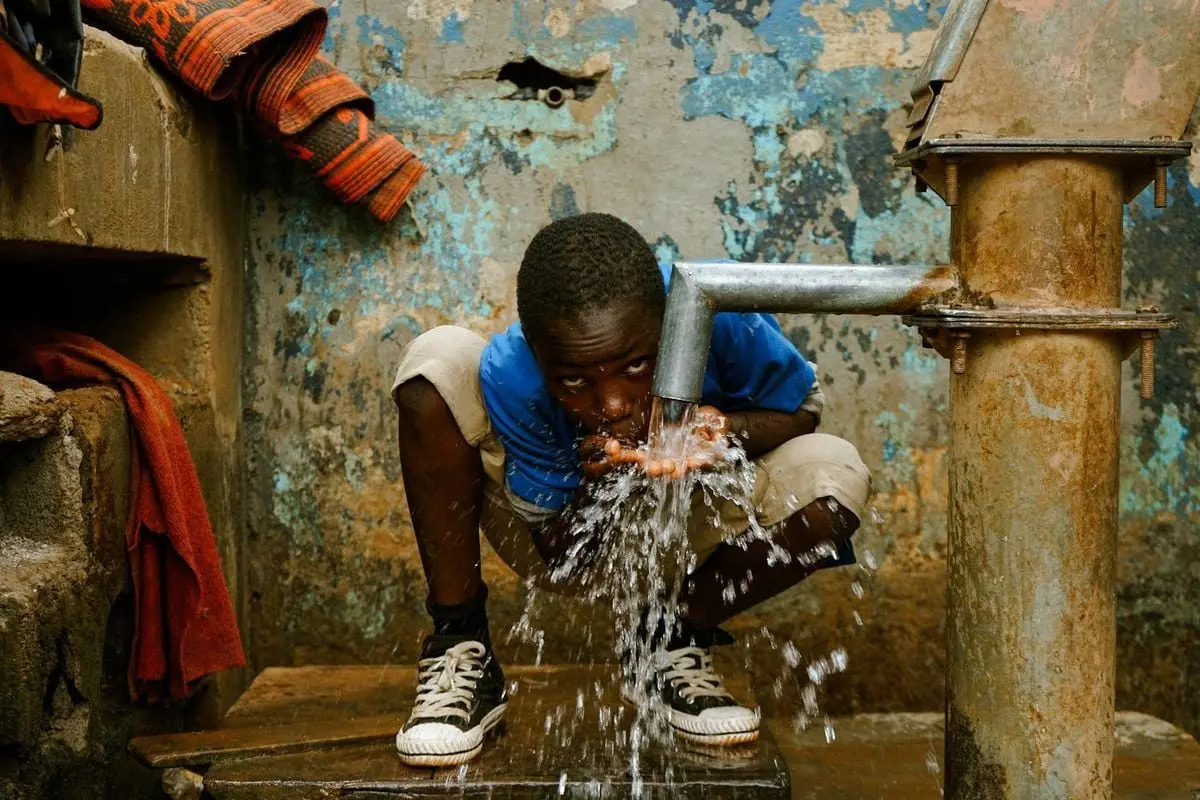 A young boy squats and drinks water from a rusty tap, emphasizing access to clean water.